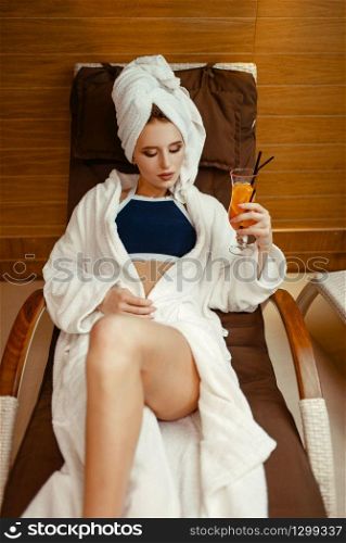 Sexy lady in robe and towel on the head relaxing with cocktail in spa chair. Relaxation leisure, healthy lifestyle, attractive woman in beauty salon. Sexy lady relaxing with cocktail in spa chair