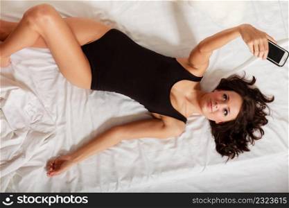 Sexy lazy girl in body underwear lying on the bed taking self photo with mobile phone. Woman relaxing lazing in the bedroom.