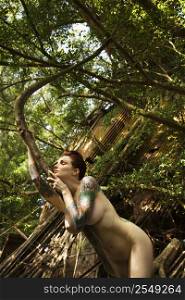 Sexy nude Caucasian tattooed woman in forest leaning on tree in Maui, Hawaii, USA.