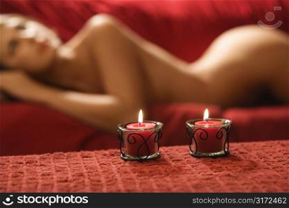 Sexy nude Caucasian young adult female lying seductively on red couch with candles.