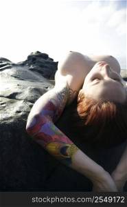 Sexy nude tattooed Caucasian woman lying on rocks overlooking Pacific Ocean in Maui, Hawaii, USA.