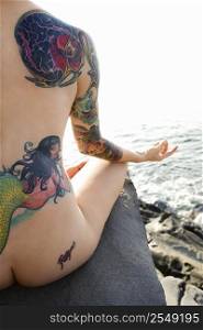 Sexy nude tattooed Caucasian woman sitting on rock meditating on beach in Maui, Hawaii, USA.