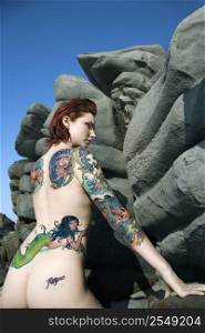 Sexy nude tattooed Caucasian woman standing by rocky formation in Maui, Hawaii, USA.