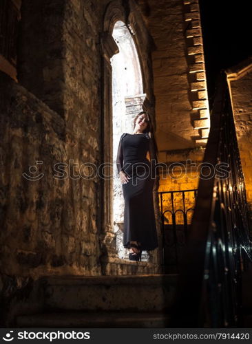 Sexy slim woman in long dress leaning against old stone arch at night