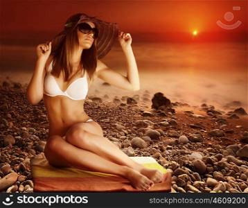 Sexy tanned model posing on the beach on red dramatic sunset background, stony coastline, fog over sea, seduction and beauty concept, summer vacation