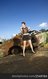 Sexy tattooed Caucasian woman standing giving middle finger in front of old rusted car in junkyard.