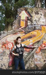 Sexy tattooed Caucasian woman standing next to wall covered in graffiti and a tree branch.