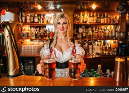 Sexy waitress holds two mugs of fresh beer at the counter in pub. Octoberfest barmaid with attractive shapes in traditional style dress
