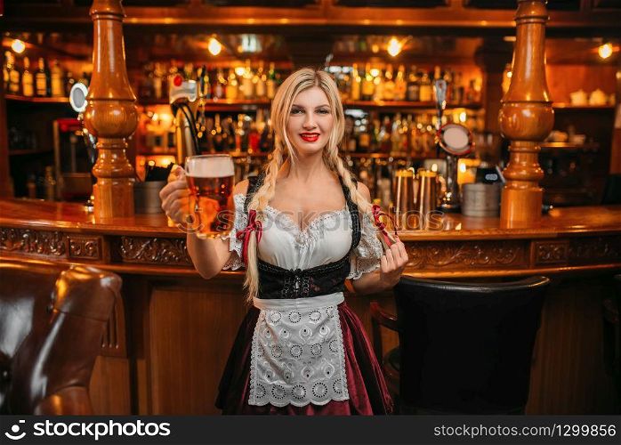 Sexy waitress holds two mugs of fresh beer in pub. Octoberfest barmaid with attractive shapes in traditional style dress. Sexy waitress holds two mugs of fresh beer in pub