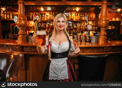 Sexy waitress holds two mugs of fresh beer in pub. Octoberfest barmaid with attractive shapes in traditional style dress. Sexy waitress holds two mugs of fresh beer in pub
