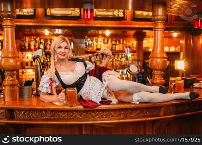 Sexy waitress in retro uniform holds a mug of fresh beer lays on counter in pub. Octoberfest barmaid with attractive shapes in traditional style dress. Sexy waitress in retro uniform holds mugs of beer