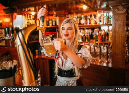 Sexy waitress pours beer into a mug at the counter in pub. Octoberfest barmaid with attractive shapes in traditional style dress