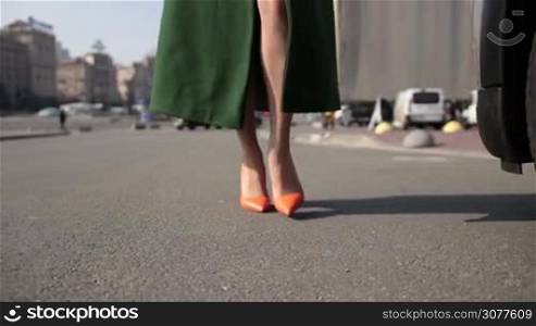 Sexy woman&acute;s legs in orange classic high heels shoes walking in the street to parked car. Low angle. Slim female legs in high heels approaching vehicle transport in parking lot and pressing car key remote control. Slow motion.