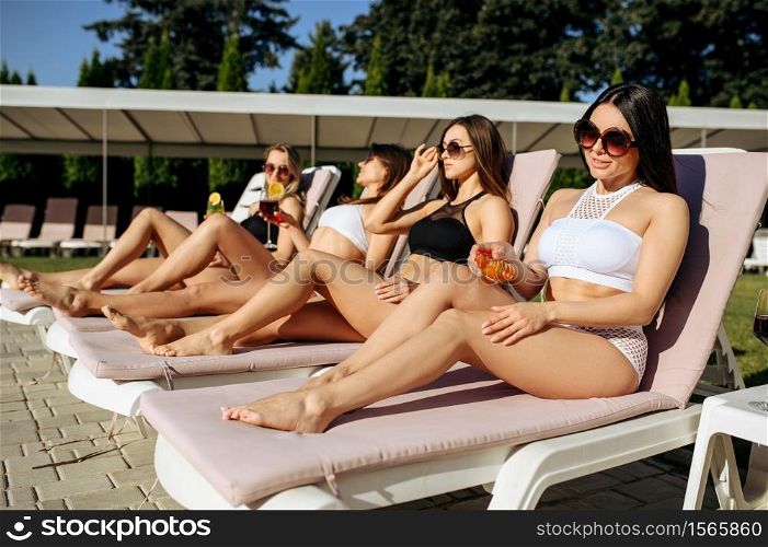Sexy woman applies tanning spray on sunbed, sunbathing. Beautiful girls relax at the poolside in sunny day, summer vacation of attractive girlfriends. Sexy woman applies tanning spray on sunbed