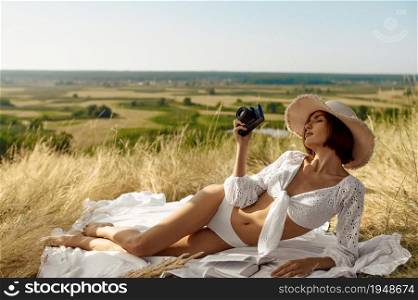 Sexy woman in lingerie and hat lying on blanket in the field. Sexy female person with slim body in underwear, lady with camera leisures on meadow, relaxation outdoors, feeling of freedom. Sexy woman in hat lying on blanket in the field