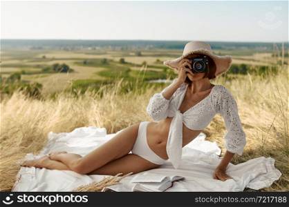 Sexy woman in lingerie and hat lying on blanket in the field. Sexy female person with slim body in underwear, lady with camera leisures on meadow, relaxation outdoors, feeling of freedom. Sexy woman in hat lying on blanket in the field