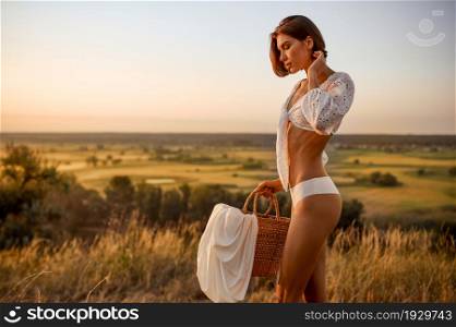 Sexy woman in white lingerie holds basket in the field. Female person with slim body in underwear leisures on meadow on sunset. Woman in white lingerie holds basket in the field