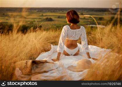 Sexy woman in white lingerie sitting on blanket in the field. Female person with slim body in underwear leisures on meadow, relaxation on sunset, feeling of freedom. Woman in white lingerie on blanket in the field