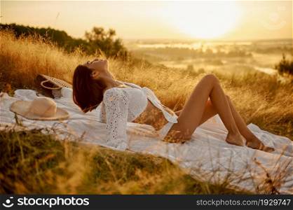 Sexy woman in white lingerie sitting on blanket in the field. Female person with slim body in underwear leisures on meadow, relaxation on sunset, feeling of freedom. Woman in white lingerie on blanket in the field