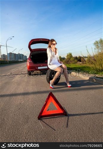 Sexy woman sitting on spare wheel against of broken car and warning sign