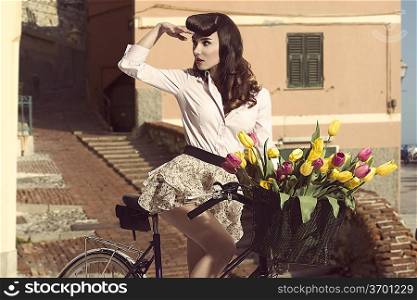 sexy woman wearing a floral skirt like vintage pin-up sitting on bicycle with some colorful flowers in the basket in old town
