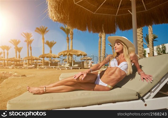 Sexy woman wearing bikini sitting on lounger under straw canopy umbrella at the beach.. Sexy woman wearing bikini sitting on lounger under straw canopy umbrella at the beach