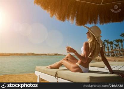 Sexy woman wearing bikini standing under straw canopy umbrella at the beach.. Sexy woman wearing bikini sitting on lounger under straw canopy umbrella at the beach holding glass with cocktail or drink