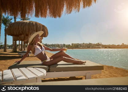 Sexy woman wearing bikini standing under straw canopy umbrella at the beach.. Sexy woman wearing bikini sitting on lounger under straw canopy umbrella at the beach holding glass with cocktail or drink