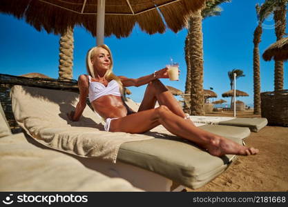 Sexy woman wearing bikini standing under straw canopy umbrella at the beach.. Sexy woman wearing bikini sitting on lounger under straw canopy umbrella at the beach holding glass with cocktail or drink