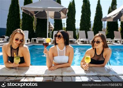 Sexy women drinks coctails in the pool outdoors. Beautiful girls in sunglasses relax at the poolside in sunny day, summer holidays of attractive girlfriends. Sexy women drinks coctails in the pool outdoors