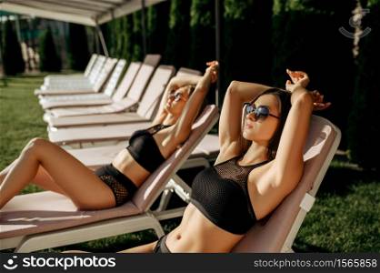 Sexy women in sunglasses sunbathing on sunbeds at the pool. Beautiful girls relax at the poolside in sunny day, summer vacation of attractive girlfriends. Sexy women in sunglasses sunbathing on sunbeds