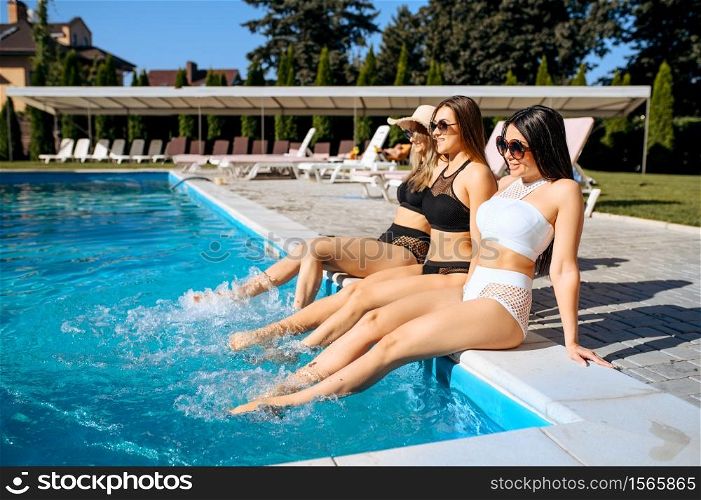 Sexy women wets their feet in the pool on resort. Beautiful girls relax at the poolside in sunny day, summer holidays of attractive girlfriends. Sexy women wets their feet in the pool on resort
