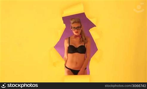 Sexy young blonde woman in a bikini wearing glasses leaning through a hole cut into yellow paper with copyspace