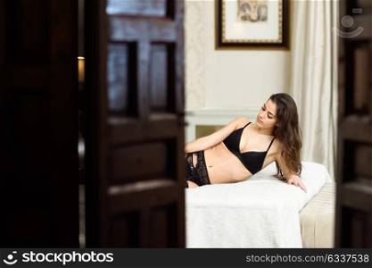 Sexy young woman in lingerie posing on the bed looking seen through a door. Brunette girl with black underwear in her bedroom