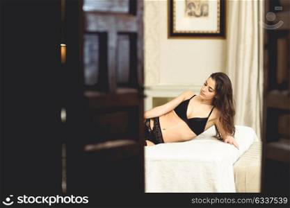 Sexy young woman in lingerie posing on the bed looking seen through a door. Brunette girl with black underwear in her bedroom