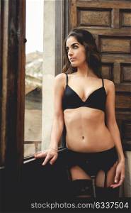 Sexy young woman in lingerie standing near a window in her bedroom. Brunette girl wearing black underwear