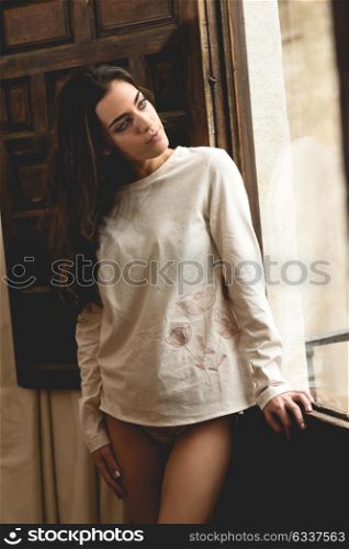 Sexy young woman in lingerie standing near a window in her bedroom. Brunette girl wearing white panties and casual t-shirt.