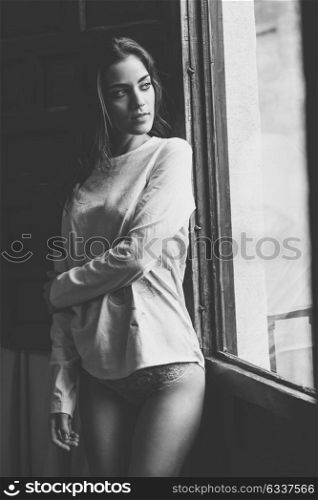Sexy young woman in lingerie standing near a window in her bedroom. Brunette girl wearing white panties and casual t-shirt. Black and white photograph.