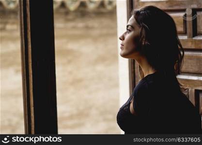 Sexy young woman in lingerie standing near a window in her bedroom. Brunette girl wearing black body and neglige. Female in underwear