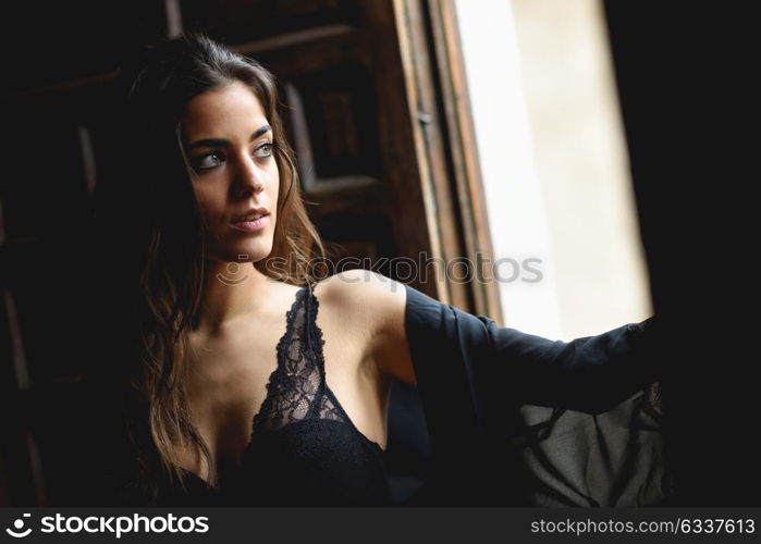 Sexy young woman in lingerie standing near a window in her bedroom. Brunette girl wearing black body and neglige. Female in underwear