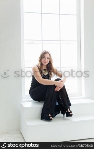 Sexy young woman in long black dress sitting on stairs at big window