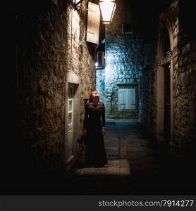 Sexy young woman in long black dress standing under lantern at old narrow street