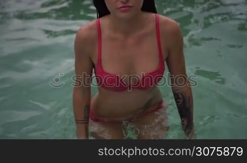 Sexy young woman wearing pink bikini with wet hair looking at camera while walking out of outdoor swimming pool at tropical resort near sea - video in slow motion