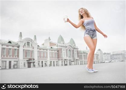 Sexy young woman with long curly hair holding a take away coffee cup, reaching out her hand and offering coffee against city background.