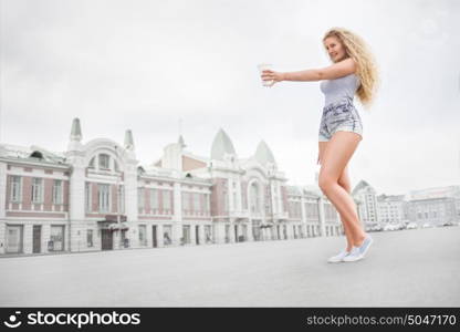 Sexy young woman with long curly hair holding a take away coffee cup, reaching out her hand and offering coffee against city background.