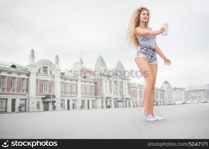 Sexy young woman with long curly hair holding a take away coffee cup, reaching out her hand and offering coffee against city background.