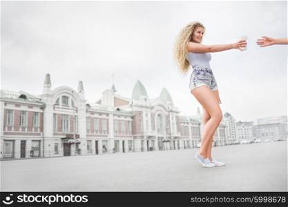 Sexy young woman with long curly hair holding a take away coffee cup, reaching out her hand and offering coffee against city background.