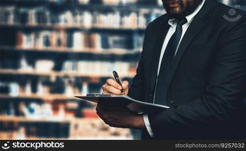 Sharp-suited businessman reviews and works on financial reports and paperwork with focused attention in his office with background of bookshelf in library. Equilibrium. Sharp-suited businessman reviews and works on financial reports. Equilibrium