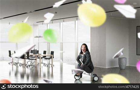 She is acting like a child. Young pretty businesswoman riding three wheeled bike in office