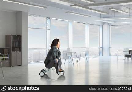 She is acting like a child. Young pretty businesswoman riding three wheeled bike in office